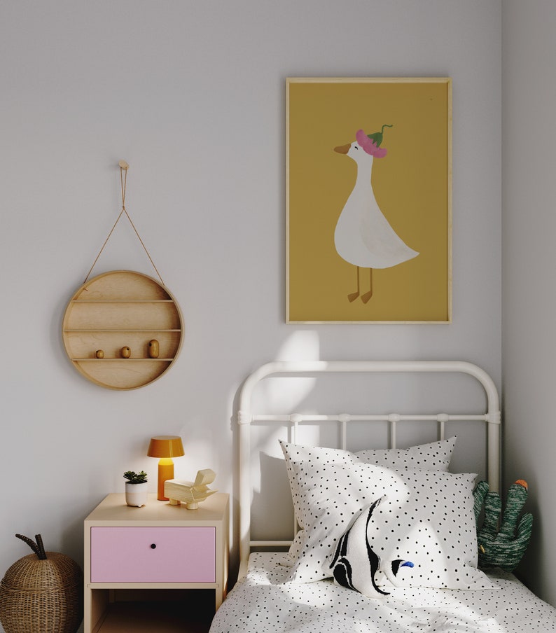 May include: A white single bed with a white metal frame and a white and black polka dot duvet cover. A white pillow with black polka dots and a black and white penguin pillow are on the bed. A green cactus plush toy is on the bed. A yellow lamp is on a pink nightstand next to the bed. A round wooden shelf with three shelves is hanging on the wall. A framed print of a white goose wearing a pink flower hat on a yellow background is hanging on the wall.