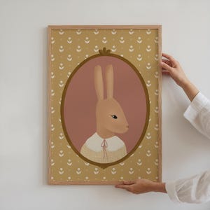 May include: A framed illustration of a brown rabbit wearing a yellow and white checkered dress with a pink background and a gold frame. The frame is surrounded by a yellow pattern of small white flowers.