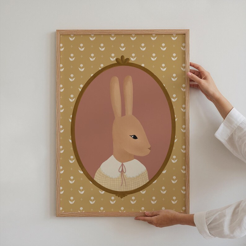 May include: A framed illustration of a brown rabbit wearing a yellow and white checkered dress with a pink background and a gold frame. The frame is surrounded by a yellow pattern of small white flowers.