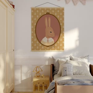 May include: A nursery room with a light brown wooden bed, a white nightstand with a lamp, and a framed print of a brown rabbit wearing a sweater. The print is hanging on a white wall with a garland of pink and white flags.