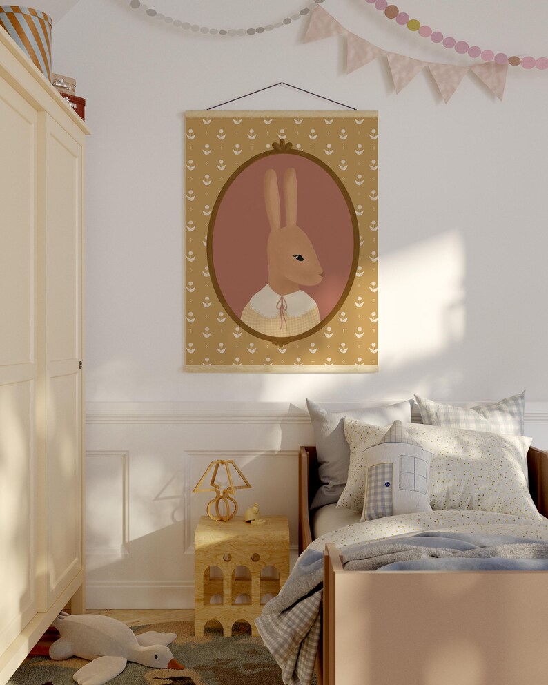 May include: A nursery room with a light brown wooden bed, a white nightstand with a lamp, and a framed print of a brown rabbit wearing a sweater. The print is hanging on a white wall with a garland of pink and white flags.