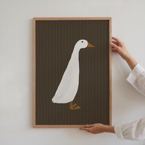 Pode incluir: Uma impress&atilde;o art&iacute;stica emoldurada com uma ilustra&ccedil;&atilde;o de um pato branco sobre um fundo castanho com riscas verticais. O pato tem bico e patas amarelas. A moldura &eacute; de madeira castanha clara.