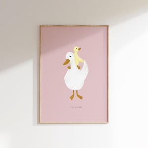 May include: A framed art print featuring a white duck carrying a yellow duckling on its back against a pink background. The text "I love you baby" is at the bottom. The artwork has a minimalist, whimsical style.