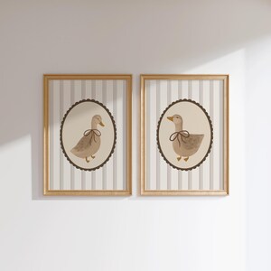 May include: Two framed prints with a brown and white striped background. Each print features a brown duck with a brown ribbon around its neck, set against a cream background inside a brown scalloped circle.