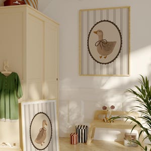 May include: Two framed prints of a brown duck with a brown bow on a white background with brown stripes. The prints are hanging on a white wall in a room with a wooden bench and a green plant.