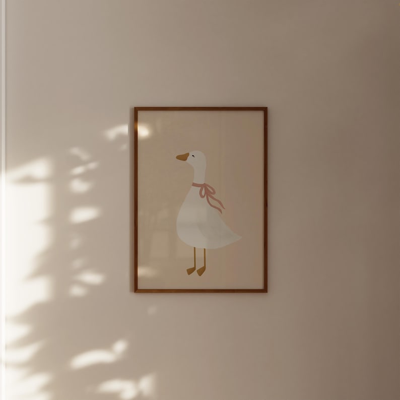 May include: A framed print featuring a whimsical illustration of a white goose with a pink ribbon around its neck. The goose has a yellow beak and feet, set against a light beige background. The frame is a warm brown color, adding a touch of rustic charm.
