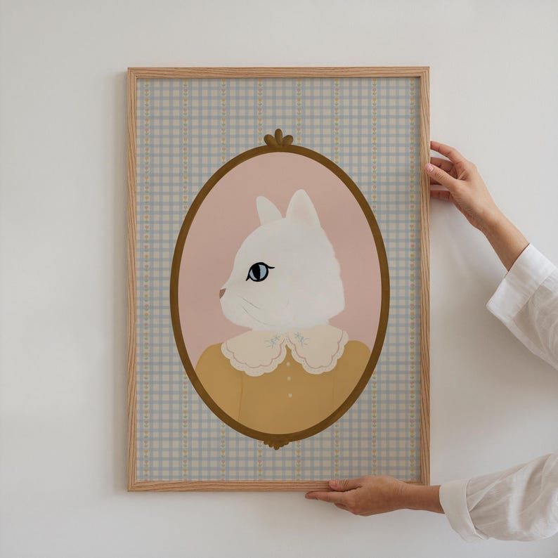 May include: A framed illustration of a white cat wearing a yellow dress with a lace collar. The cat is looking to the left and is set against a light blue and white gingham background.