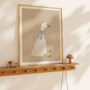 May include: A framed illustration of a white goose with a blue bow sitting on a light brown background. The frame is a light wood color. The illustration is on a wooden shelf with a small mushroom figurine on the right side of the shelf. A small toy camera hangs from a hook on the shelf.