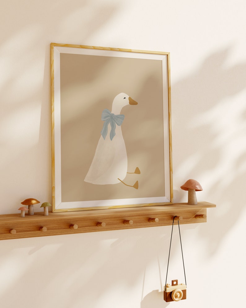 May include: A framed illustration of a white goose with a blue bow sitting on a light brown background. The frame is a light wood color. The illustration is on a wooden shelf with a small mushroom figurine on the right side of the shelf. A small toy camera hangs from a hook on the shelf.