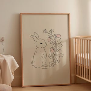 May include: A framed print of a white rabbit standing next to a pink strawberry plant with green leaves and flowers. The print is on a cream-colored background.