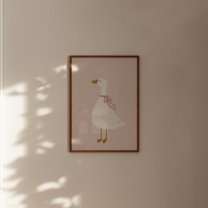 May include: A framed print featuring a whimsical illustration of a white goose with a pink ribbon around its neck. The goose has a yellow beak and legs, set against a soft pink background. The frame is a warm brown color.