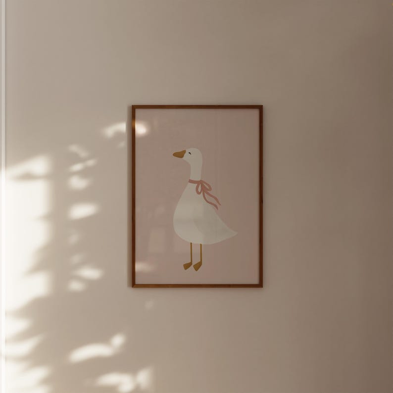 May include: A framed print featuring a whimsical illustration of a white goose with a pink ribbon around its neck. The goose has a yellow beak and legs, set against a soft pink background. The frame is a warm brown color.