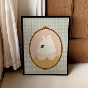 May include: A white cat with blue eyes wearing a yellow dress with a white lace collar is framed in a gold oval on a blue and white gingham background.