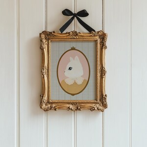 May include: A framed illustration of a white cat with blue eyes wearing a yellow collar with a white lace trim. The cat is in a pink oval frame on a blue and white gingham background.