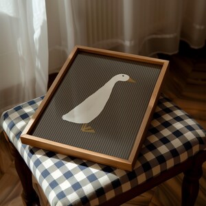Pode incluir: Uma impress&atilde;o art&iacute;stica emoldurada com uma ilustra&ccedil;&atilde;o de um ganso branco sobre um fundo listrado cinza. A obra de arte est&aacute; emoldurada em uma moldura de madeira natural. A impress&atilde;o &eacute; exibida em um banquinho estofado xadrez azul e branco, criando uma est&eacute;tica simples e moderna.