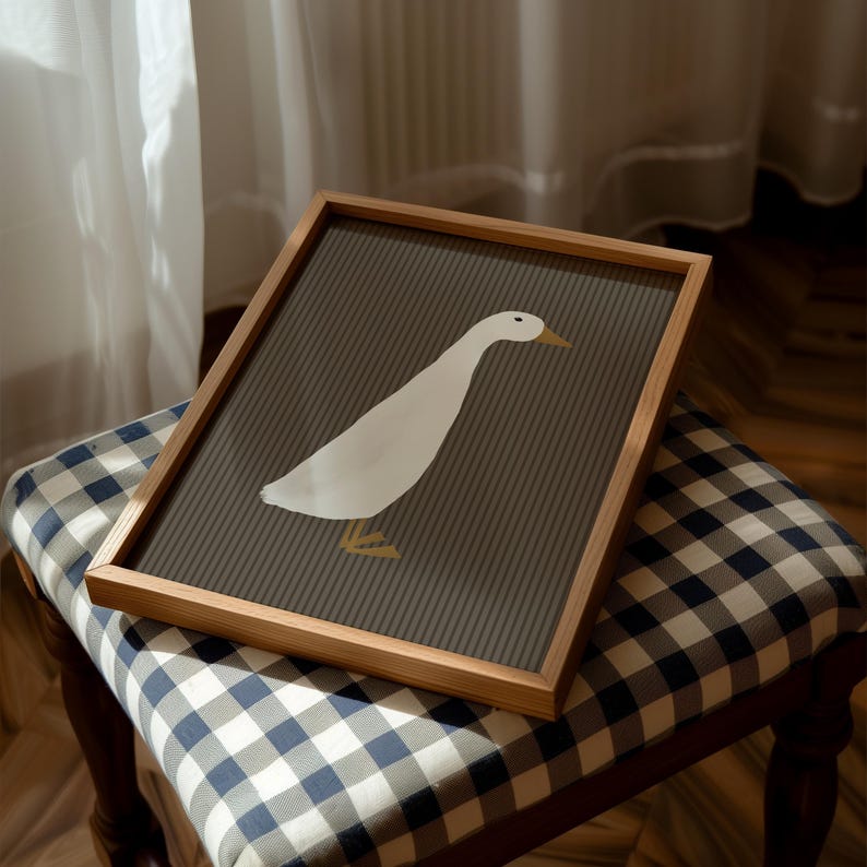 Pode incluir: Uma impress&atilde;o art&iacute;stica emoldurada com uma ilustra&ccedil;&atilde;o de um ganso branco sobre um fundo listrado cinza. A obra de arte est&aacute; emoldurada em uma moldura de madeira natural. A impress&atilde;o &eacute; exibida em um banquinho estofado xadrez azul e branco, criando uma est&eacute;tica simples e moderna.