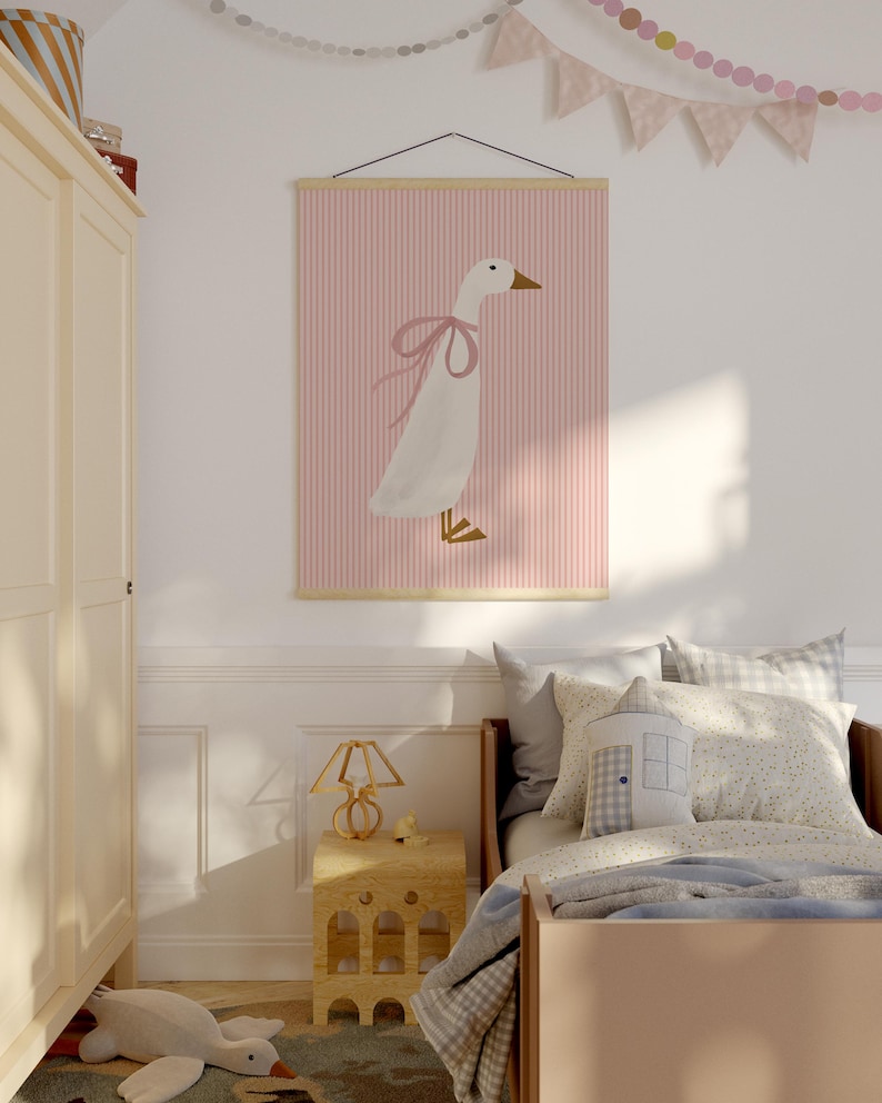 May include: A pink and white striped poster with a white goose wearing a pink bow. The poster is hanging on a white wall above a bed with a light blue blanket and a pillow with a house design.
