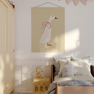 May include: A white goose with a pink bow on its neck is featured on a yellow and white striped poster hanging on a white wall. The poster is framed with a wooden frame.