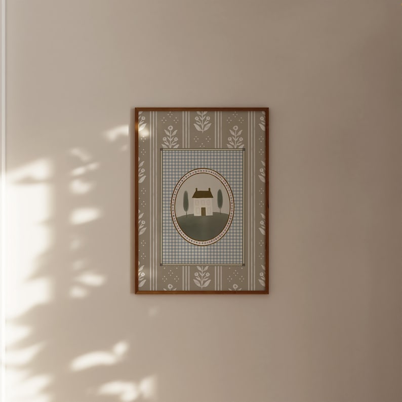 May include: A framed print with a light blue and white checkered background. The print features a white house with a brown roof and two green trees in front of it. The house is surrounded by a brown circle with a white border.
