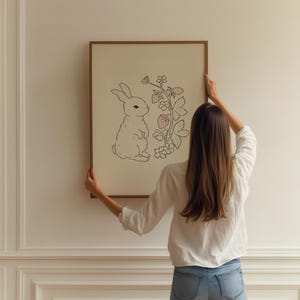 May include: A person hangs a framed illustration of a white rabbit standing next to a pink strawberry plant on a white wall.