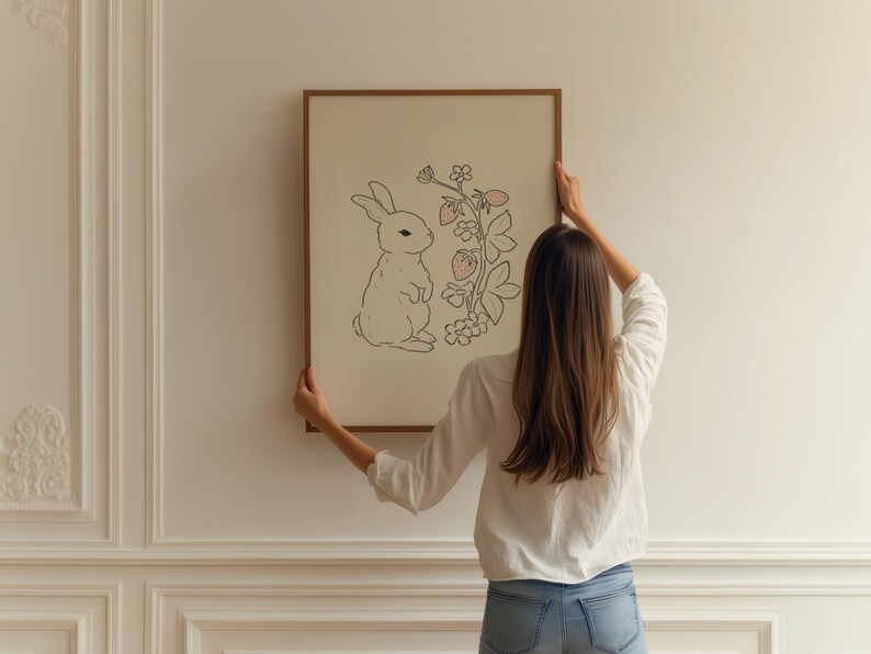 May include: A person hangs a framed illustration of a white rabbit standing next to a pink strawberry plant on a white wall.