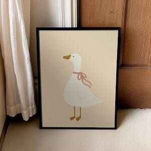 May include: Framed art print featuring a whimsical illustration of a white goose with a pink ribbon around its neck. The goose has a yellow beak and feet, set against a cream-colored background. The artwork is enclosed in a black frame.