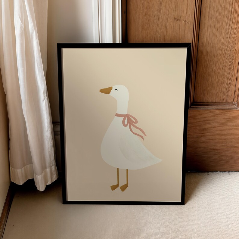 May include: Framed art print featuring a whimsical illustration of a white goose with a pink ribbon around its neck. The goose has a yellow beak and feet, set against a cream-colored background. The artwork is enclosed in a black frame.