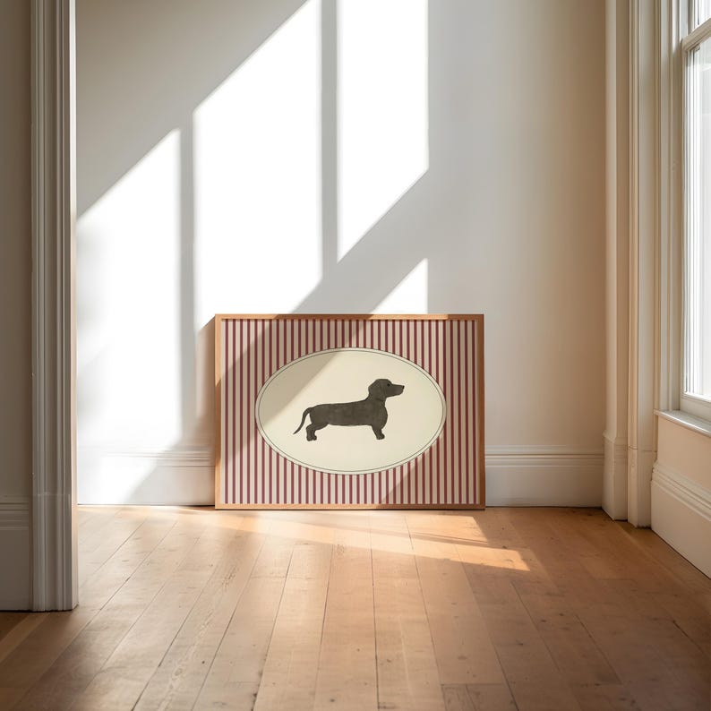 May include: A framed artwork featuring a dachshund silhouette within an oval on a striped background. The artwork is in a wooden frame and rests on a wooden floor. The background is a white wall with sunlight.