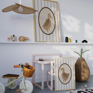 May include: Two framed prints of a brown duck with a brown bow on a white background with gray stripes. The prints are hanging on a white wall above a shelf with a wooden toy and a wicker basket. A pink table and chair are in the foreground with a wooden toy boat on the table. A wicker basket with a green plant is in the background.