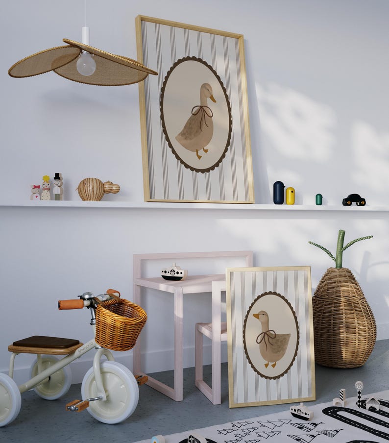 May include: Two framed prints of a brown duck with a brown bow on a white background with gray stripes. The prints are hanging on a white wall above a shelf with a wooden toy and a wicker basket. A pink table and chair are in the foreground with a wooden toy boat on the table. A wicker basket with a green plant is in the background.