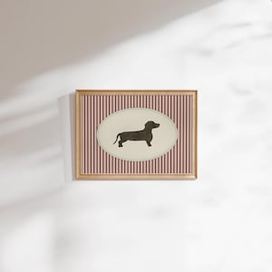 May include: A framed print featuring a black dachshund silhouette on an oval cream background. The oval is set against a backdrop of vertical red and white stripes. The frame is a light wood color.