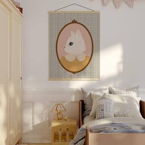 May include: A white cat with blue eyes in a yellow dress is framed in a brown oval and displayed on a blue and white checkered wall tapestry. The tapestry is hanging on a wooden rod.