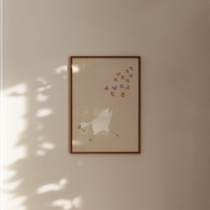 May include: A framed print of a white goose with a brown twig in its beak, flying towards a flock of purple and orange butterflies. The print has a light brown background and a wooden frame.