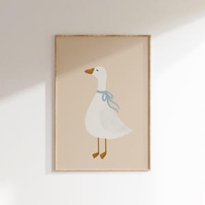 Può includere: Stampa incorniciata con un'illustrazione di un'oca bianca con un nastro blu, becco e zampe gialle. L'immagine è su sfondo beige, in una cornice di legno marrone chiaro. Adatta per una cameretta.