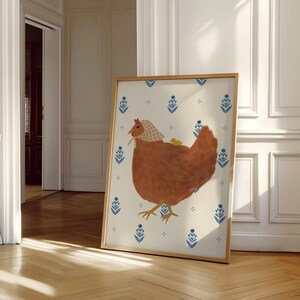 Puede incluir: Una ilustraci&oacute;n enmarcada de una gallina marr&oacute;n con un pollito amarillo en la espalda. La gallina lleva una capucha con estampado blanco y rojo. El fondo es de color crema con un patr&oacute;n floral azul y blanco repetido.