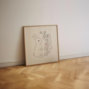 May include: A framed print of a white rabbit standing next to a pink and green strawberry plant with leaves and flowers. The print is on a cream-colored background.