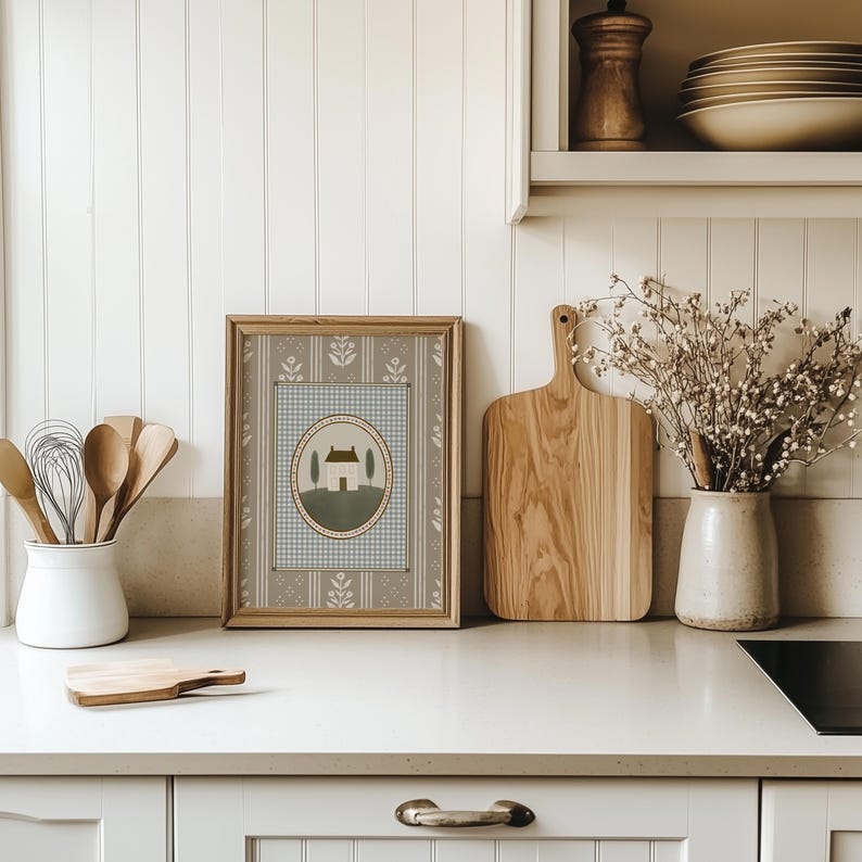 May include: A framed print of a house with two trees in a circle on a blue and white checkered background. The frame is brown wood and the print is on a beige background with a floral pattern.