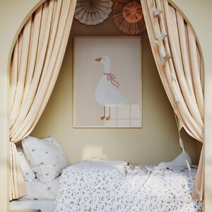 May include: A children's bedroom scene featuring a framed print of a white goose with a pink bow. The room has a bed with floral bedding, pillows, and a small wicker chest. Soft peach-colored curtains frame the bed, and decorative paper fans adorn the wall.