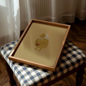 Puede incluir: Una ilustraci&oacute;n enmarcada de un pollito amarillo con un pa&ntilde;uelo estampado en blanco y azul. El pollito sostiene un peque&ntilde;o cubo rojo y est&aacute; de pie sobre un fondo marr&oacute;n claro.