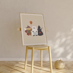 Puede incluir: Una ilustraci&oacute;n enmarcada de tres personajes disfrazados: un zorro, un fantasma y un gato negro. El zorro lleva un disfraz marr&oacute;n y naranja con un cubo de calabaza. El fantasma lleva una s&aacute;bana blanca con un patr&oacute;n de rayas azules y blancas. El gato negro lleva un disfraz negro con un cubo de calabaza. La ilustraci&oacute;n est&aacute; sobre un fondo beige y est&aacute; enmarcada en un marco de madera claro.