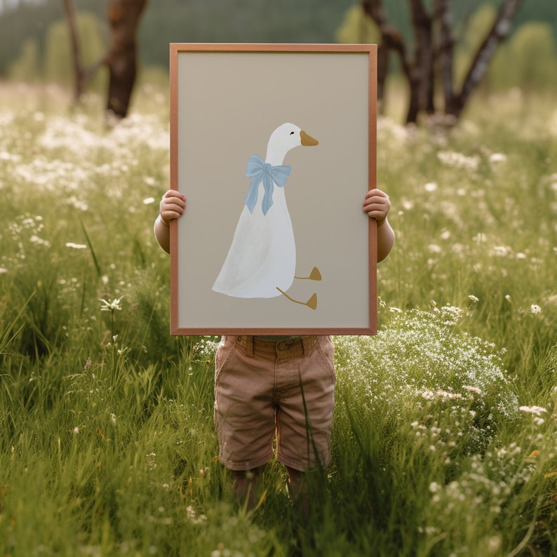 May include: A child holds a framed print of a white goose with a blue bow. The goose is sitting on a beige background.