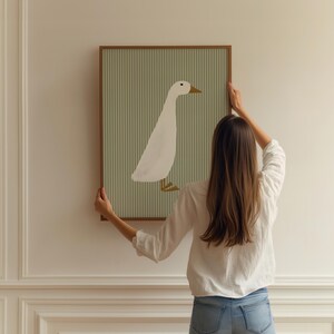 May include: A woman hangs a framed print of a white duck on a light green striped background on a white wall.