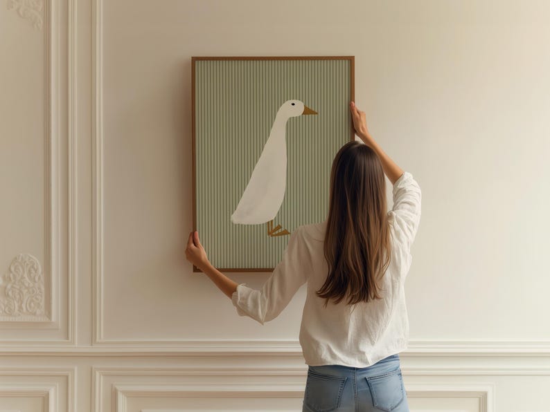 May include: A woman hangs a framed print of a white duck on a light green striped background on a white wall.
