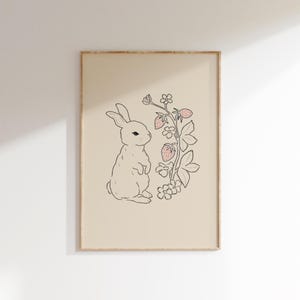 May include: A black and white line drawing of a rabbit standing next to a plant with pink strawberries. The background is a light beige color.