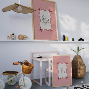 May include: Two framed prints with pink and white striped backgrounds and a black and white illustration of a duck inside a red and white frame. The prints are hanging on a white wall above a pink table and chair. A wooden toy tricycle with a wicker basket is in the foreground.