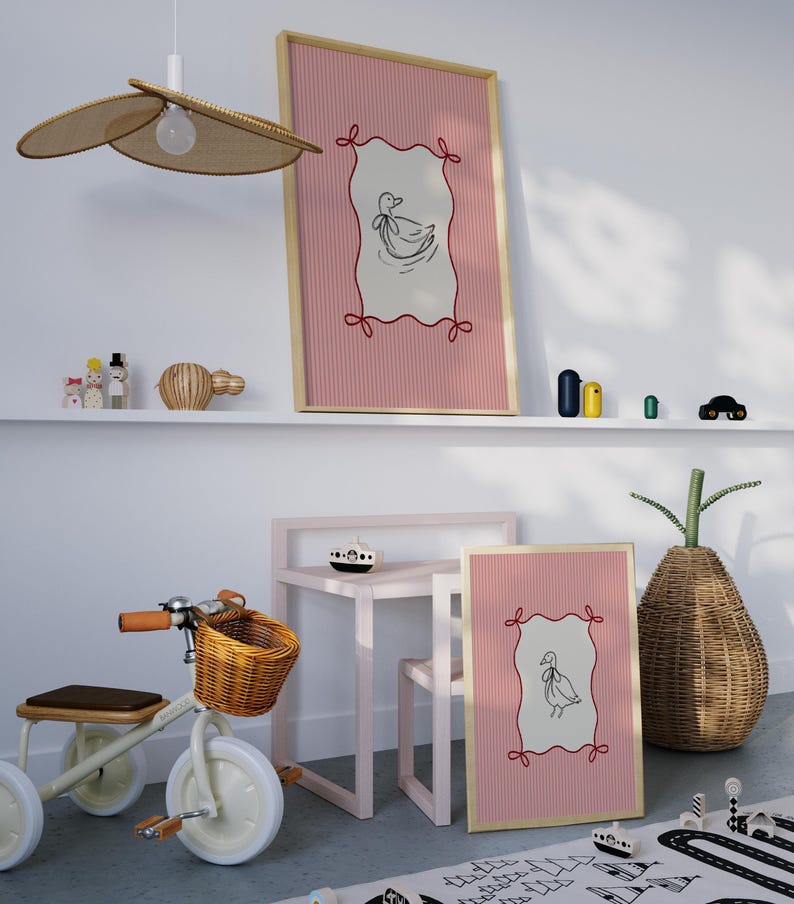 May include: Two framed prints with pink and white striped backgrounds and a black and white illustration of a duck inside a red and white frame. The prints are hanging on a white wall above a pink table and chair. A wooden toy tricycle with a wicker basket is in the foreground.
