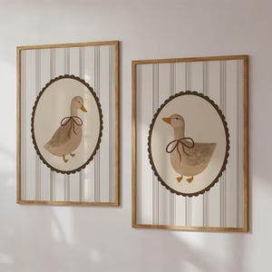May include: Two framed prints with a striped background and a brown circular border. Each print features a watercolor illustration of a duck with a brown bow.
