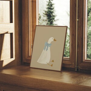 May include: A framed illustration of a white goose with a blue bow sitting on a light brown background. The frame is sitting on a wooden surface.