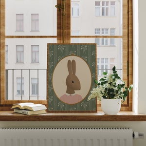 May include: A green and white floral print framed illustration of a brown rabbit wearing a pink shirt. The rabbit is looking to the left. The frame is on a wooden shelf with a book and a potted plant.