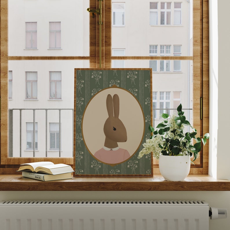 May include: A green and white floral print framed illustration of a brown rabbit wearing a pink shirt. The rabbit is looking to the left. The frame is on a wooden shelf with a book and a potted plant.